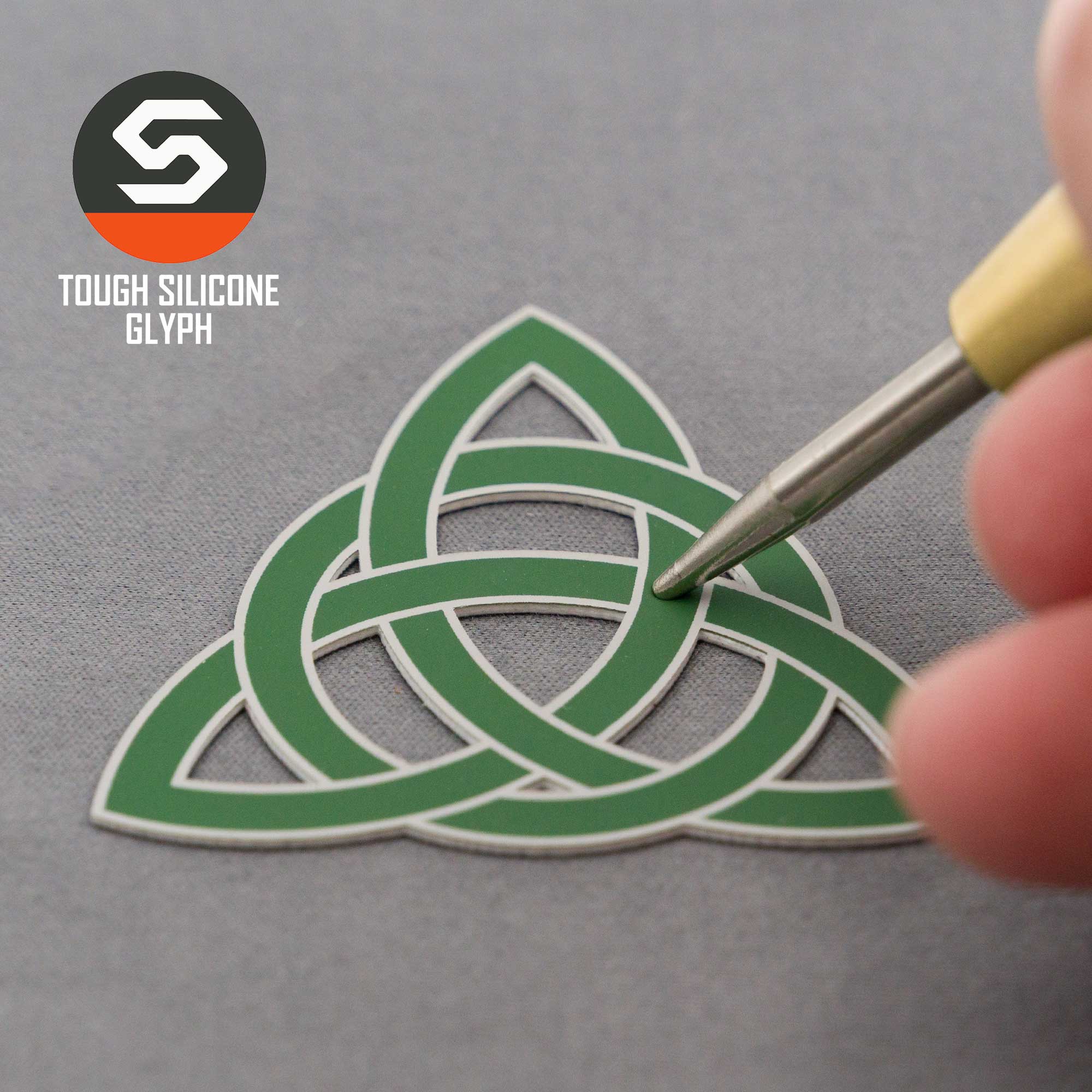 A hand uses a tool to touch a green silicone Triquetra Trinity Knot on a gray surface. The Eternal Glyph logo and the text "Triquetra Trinity Knot (Silicone) - Flexfit® Mesh Trucker Cap" appear in the top left corner.