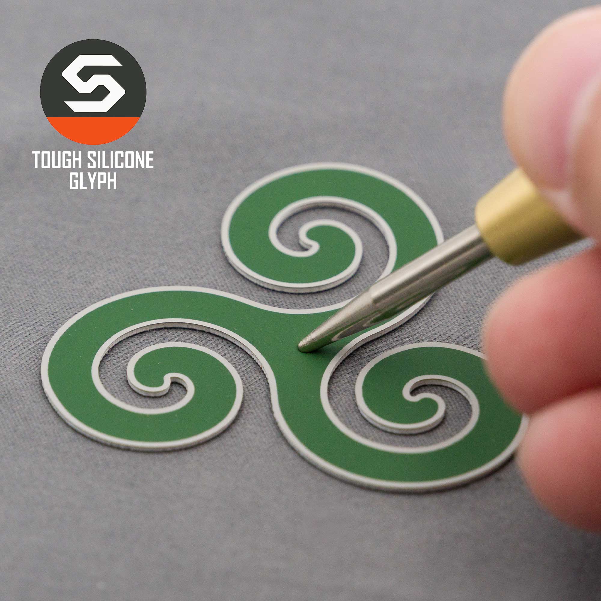 A hand adjusts an Eternal Glyph Flexfit® Mesh Trucker Cap featuring a green silicone Triskelion symbol with white outlines, inspired by Celtic art. The cap sits on a gray surface, its Tough Silicone Glyph logo in the top left corner.