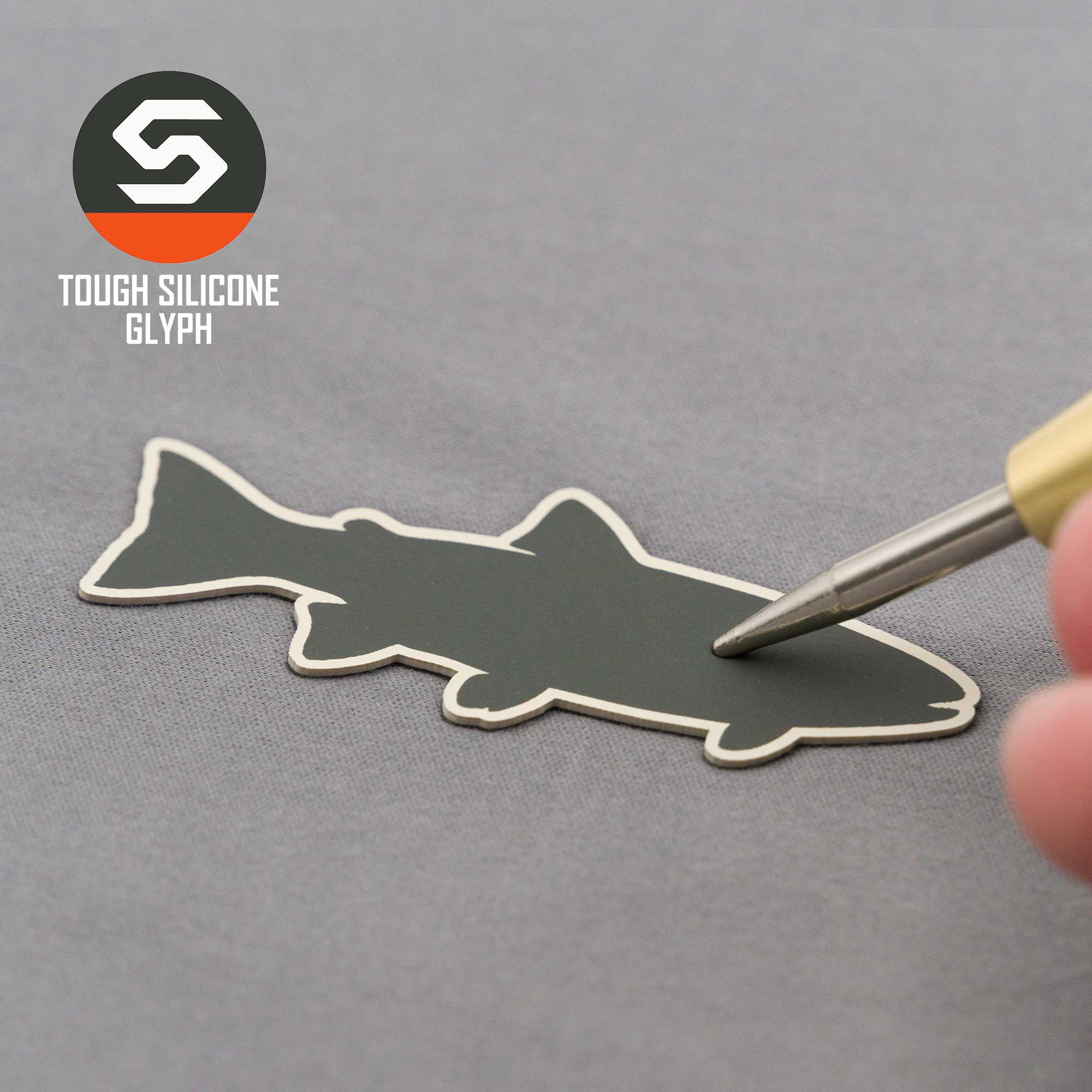 A hand touches a dark fish-shaped silicone patch on a gray surface, evoking trout fishing. The top left shows the Eternal Glyph logo: "Trout Fishing (Silicone) - Flexfit® Flat Bill Premium Fitted Cap" with a stylized S symbol.