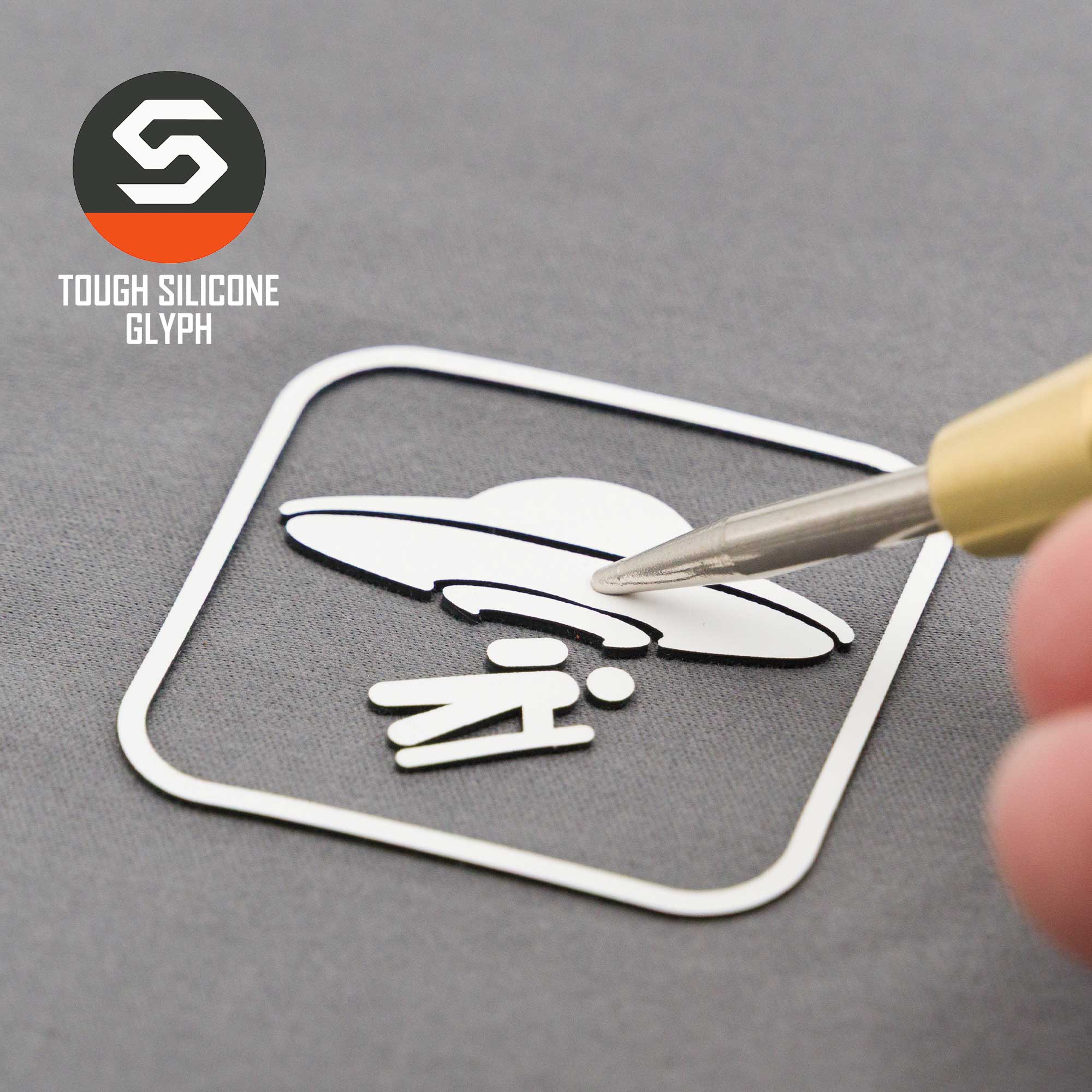 A close-up shows a hand applying the Eternal Glyph UFO Area Trail Sign (Silicone) sticker, featuring a UFO beaming up a person, onto a gray surface. The TOUGH SILICONE GLYPH logo appears in the top left corner.