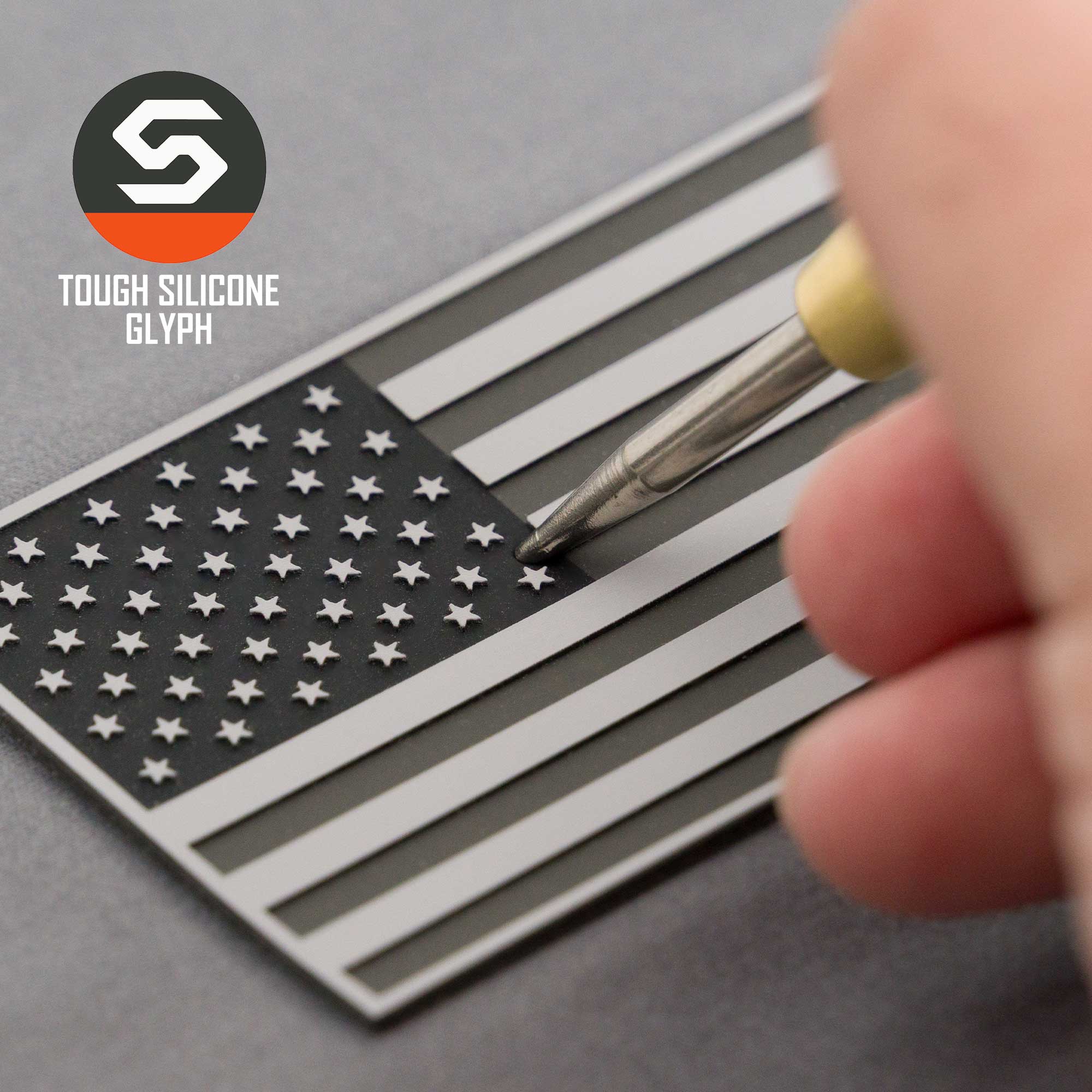 A hand presses a tool onto the grayscale American Flag, Monotone (Double Layer Silicone) patch on an Eternal Glyph Flexfit® Mesh Trucker Cap. "TOUGH SILICONE GLYPH" reflects tactical patriotism beside the top left logo.