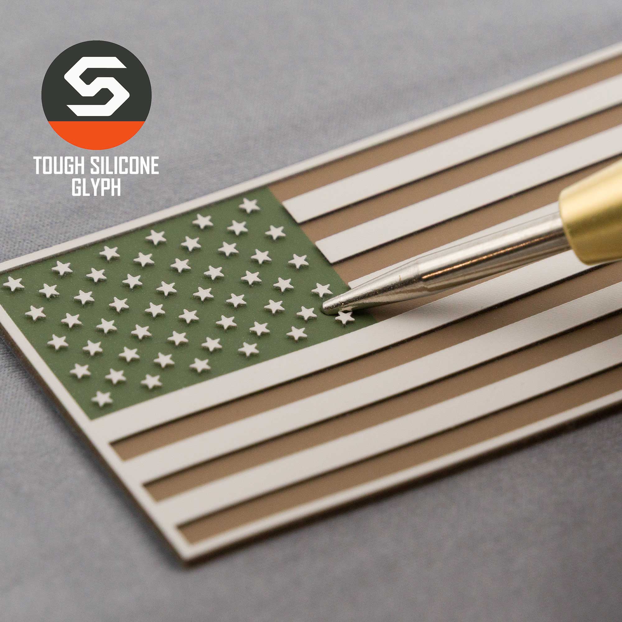A pen points at an Eternal Glyph American Flag, Camo (Double Layer Silicone) patch in olive, tan, and white on a gray surface. The Tough Silicone Glyph logo at the top left evokes rugged outdoor gear.