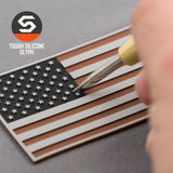 A hand uses a tool to press on a raised star atop the Eternal Glyph American Flag, Coyote Black (Double Layer Silicone) Flexfit® Mesh Trucker Cap, set on gray. The Tough Silicone Glyph logo appears in the top left, evoking military-grade durability.