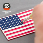 A hand works on an Eternal Glyph American Flag Flexfit® Cool & Dry Cap, featuring a double-layer silicone red, white, and blue patch. The patriotic patch and brand logo are clearly visible in the scene.