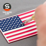 A hand works on an Eternal Glyph American Flag Flexfit® Cool & Dry Cap, featuring a double-layer silicone red, white, and blue patch. The patriotic patch and brand logo are clearly visible in the scene.