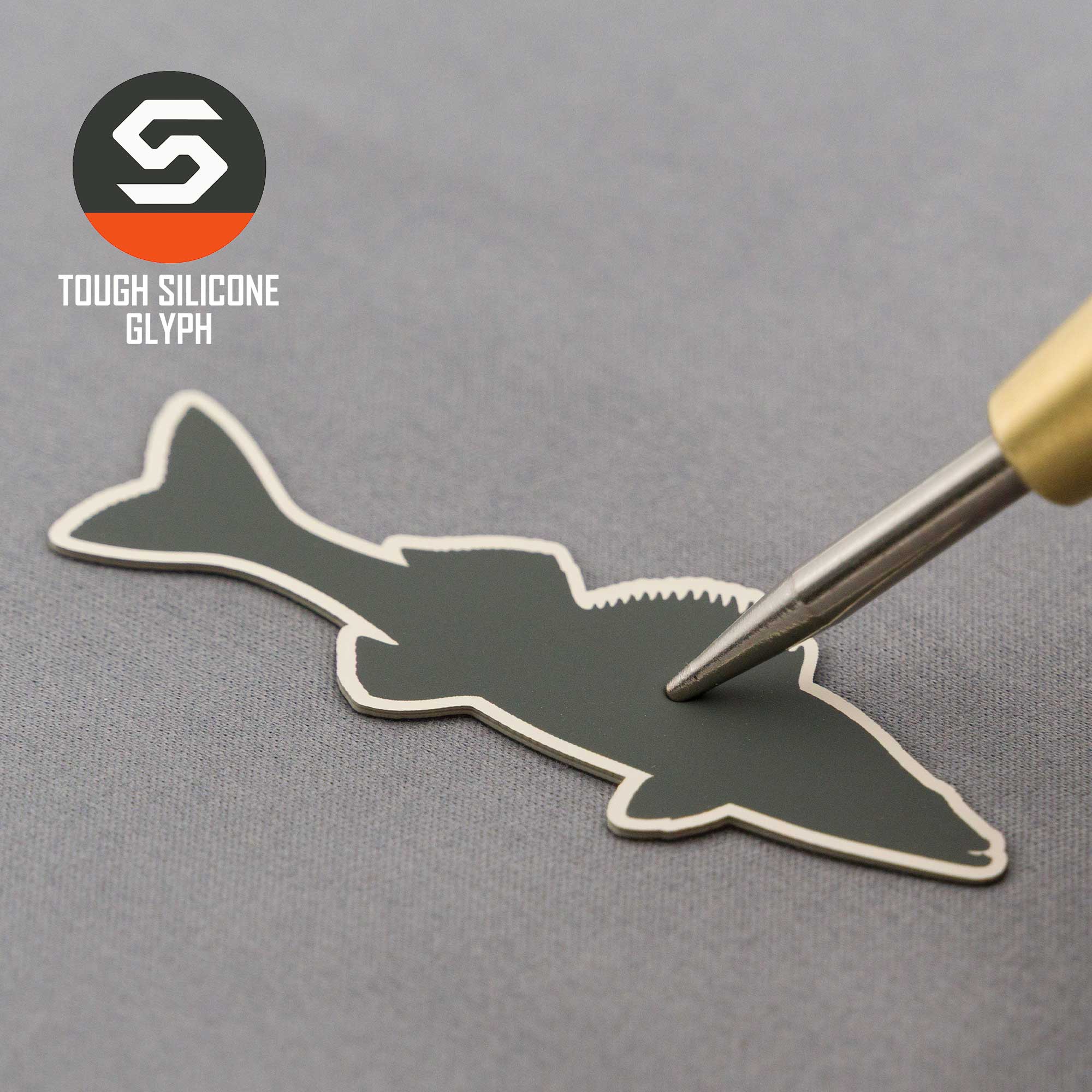 A Flexfit® Mesh Trucker Cap by Eternal Glyph, featuring a Walleye Fishing silicone silhouette patch, is shown as a metal tool pokes the patch on a gray surface with the Eternal Glyph logo in the corner.