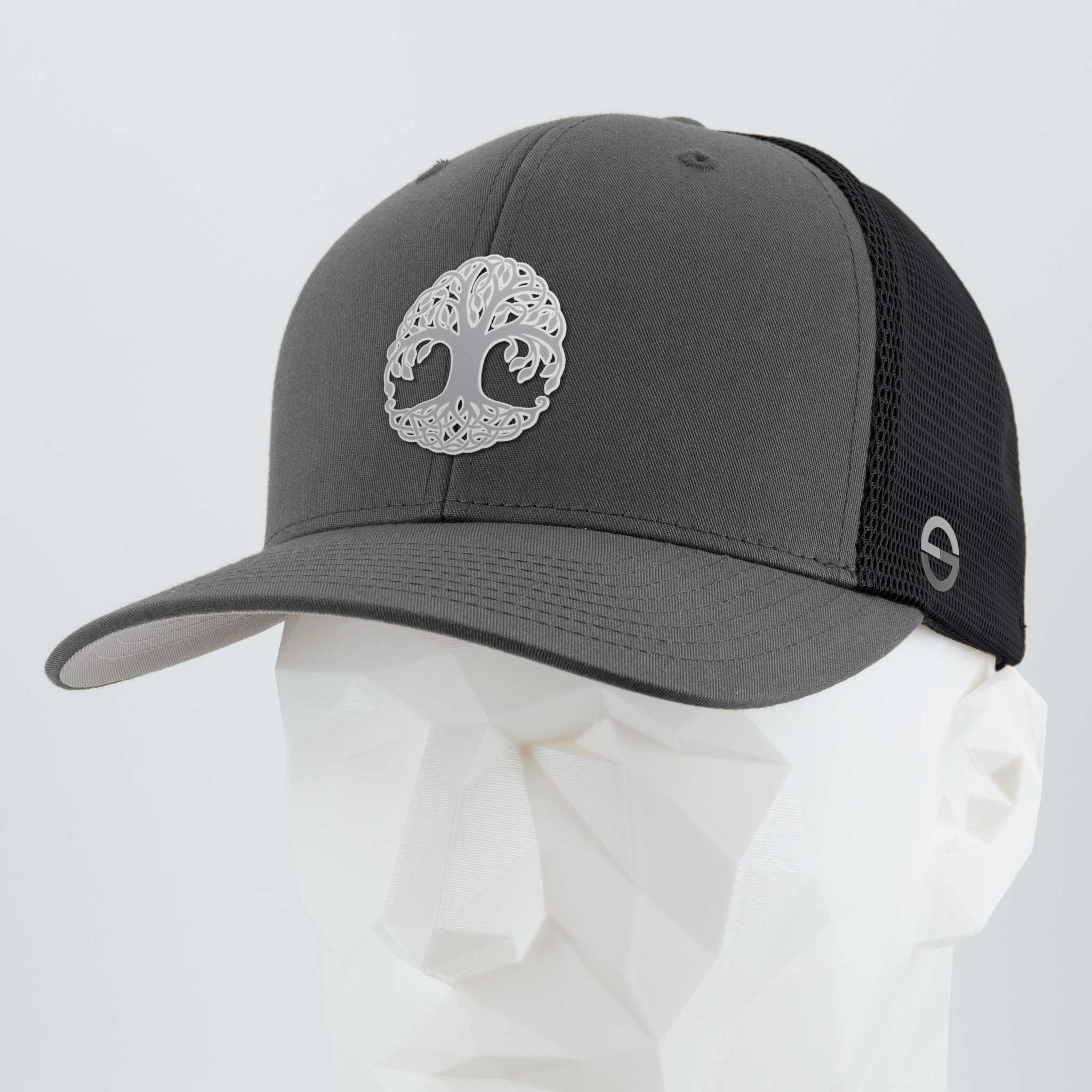 The Eternal Glyph Flexfit® Mesh Trucker Cap features a metallic silver silicone Tree of Life (Yggdrasil) symbol on gray and black mesh, shown on a white geometric mannequin against a plain light background.