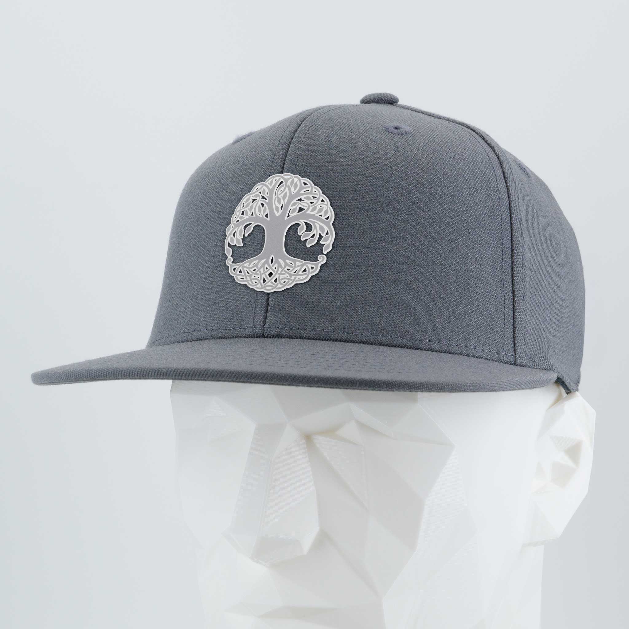 The Eternal Glyph Tree of Life, Yggdrasil Flexfit® Flat Bill Cap features a metallic silver silicone emblem and a gray design, shown on a white geometric mannequin head against a light background.
