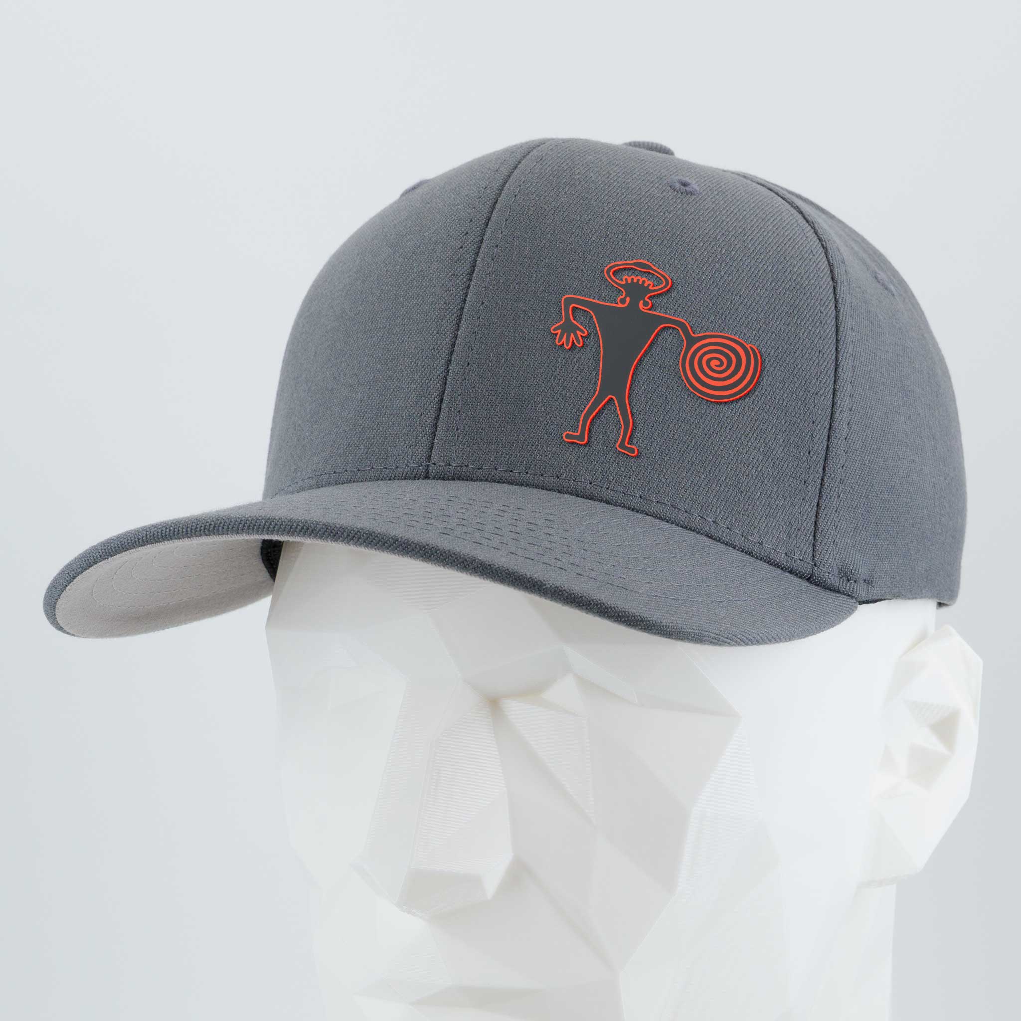 The Eternal Glyph Spiral Warrior Petroglyph (Silicone) Flexfit® Premium Wool Blend Cap in dark gray features a red-orange abstract design inspired by an ancient Moab, Utah carving. Shown on a faceted white mannequin head.